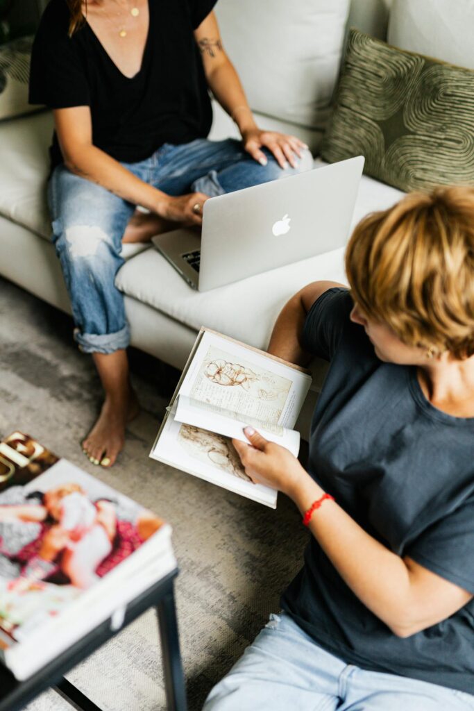 Two college age girls studying. One has a book open. The other has a laptop open. The book appears to be full of anatomy like diagrams.